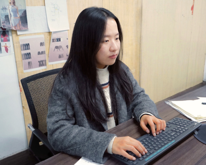 The image shows a person sitting at a desk and typing on a keyboard. The person is wearing a gray sweater and has long dark hair. The face of the person is blurred. The desk has some papers and a mouse on it. The background includes a wall with several pieces of paper pinned to it, which appear to have various designs or sketches on them.